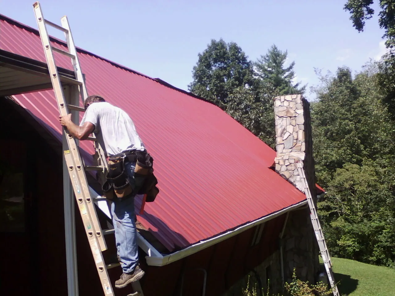 Roofer on a ladder beside a red metal roof and stone chimney — Soffit & Fascia Repair by Roofing Brothers in Sherwood